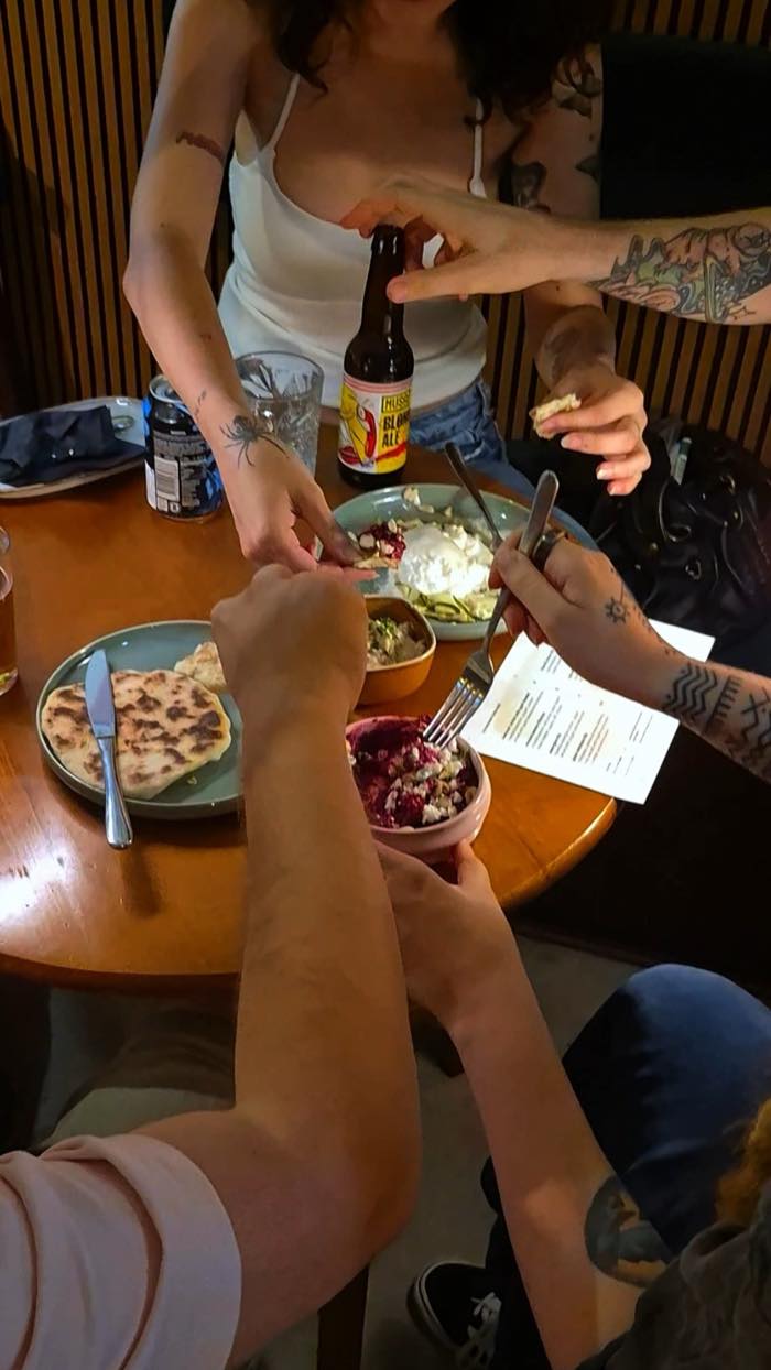 Mesa partilhada no DOM: mãos a servir dips, pão achatado e cerveja.